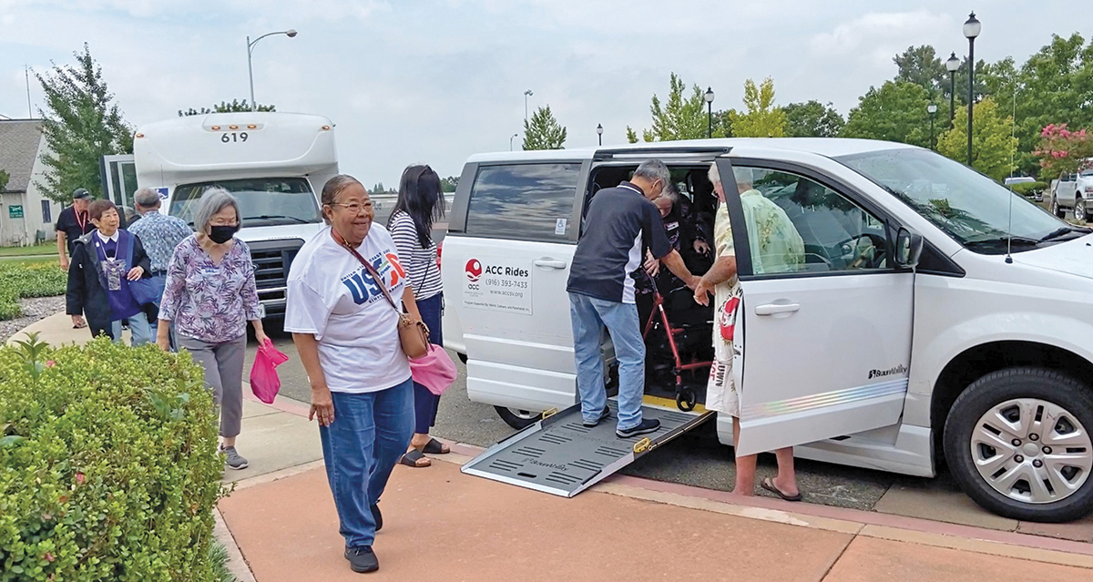 A group of people walking to or from a pair of white passenger vans with one person being helped into one. 