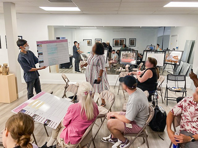 Group of people sitting and listening to a presenter at the front of a room holding a poster.