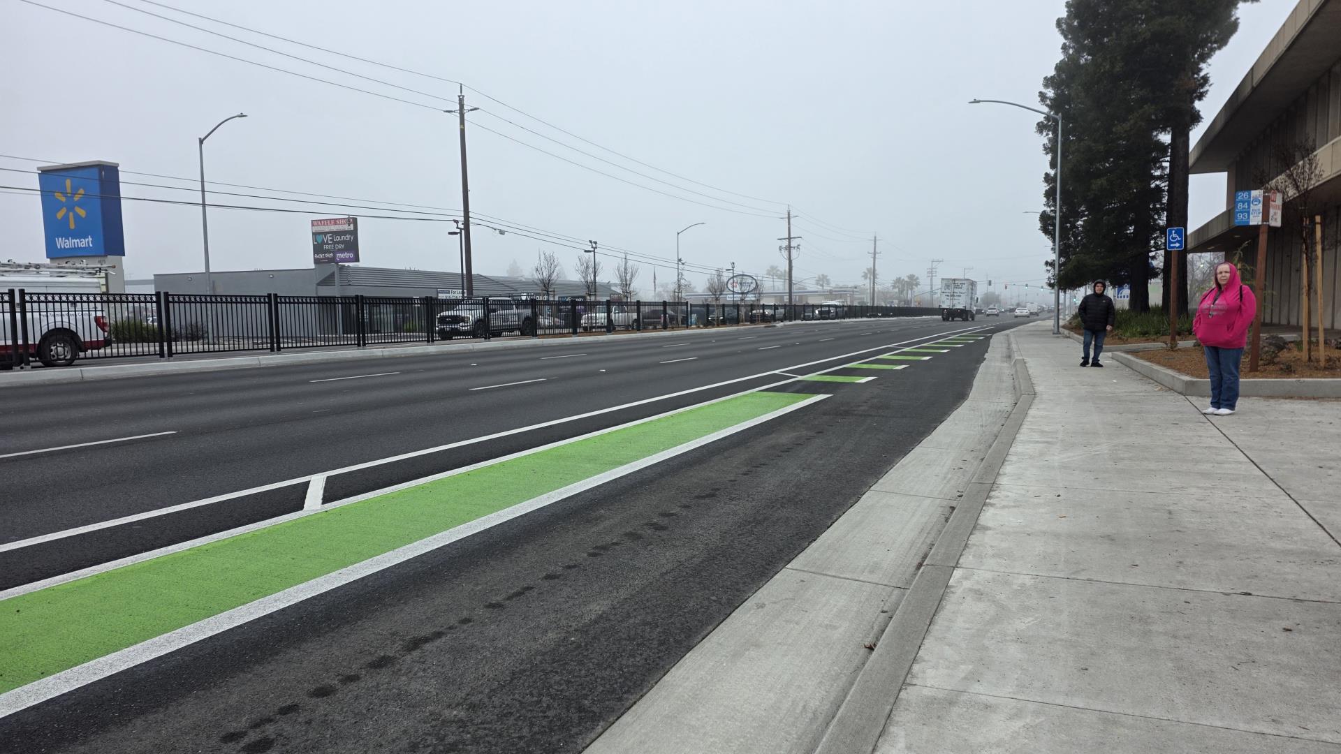 Looking down Watt Avenue with bike lanes painted green, new sidewalks and two people waiting for the bus in the distance